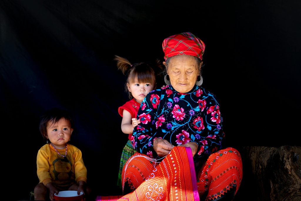 An old lady from Eastern India designing a red colored cloth, sitting with two young kids