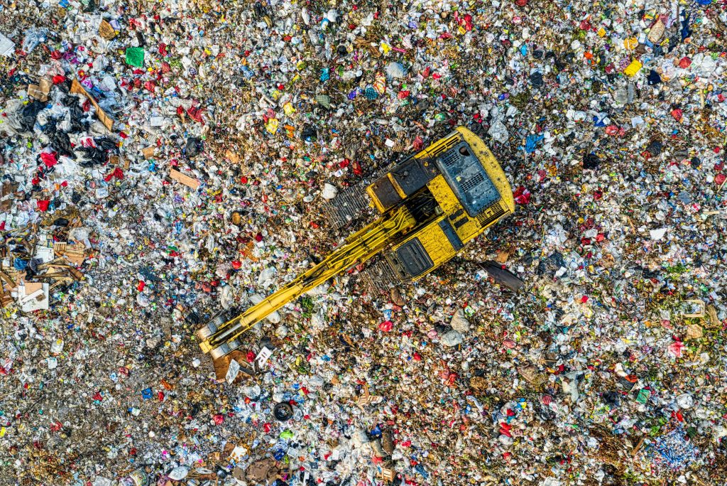 A landfill of discarded clothes, plastic items, and other things, with a dump truck in the middle, polluting the environment.