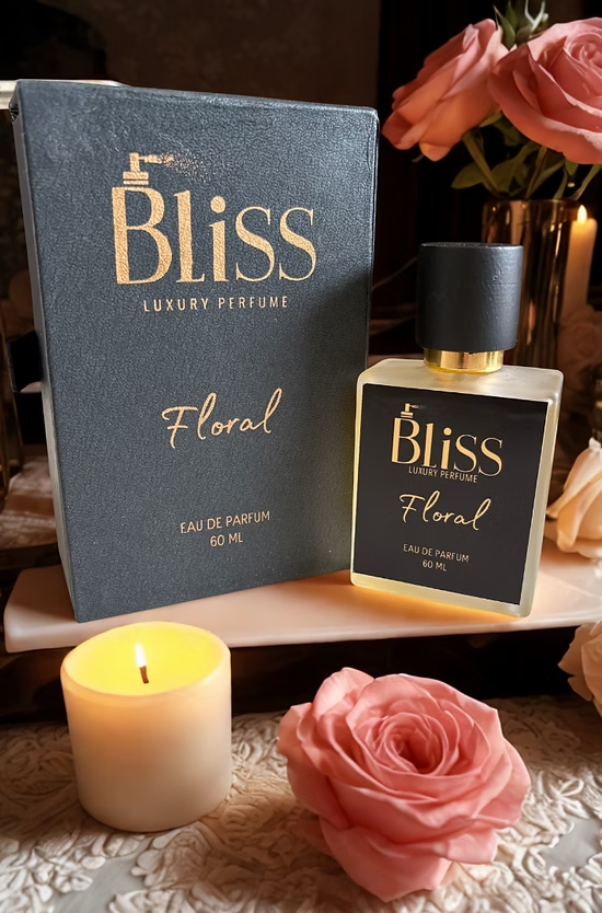 A bottle of Bliss Floral Eau De Parfum placed on a desk with roses in the background and a candle lit in the foreground with a pink rose besides it