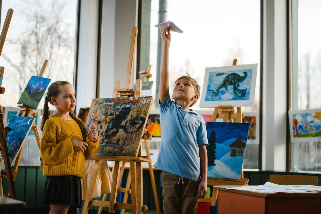 Two children in a classroom - a boy flying a paper airplane while a girl watches, with easels and colorful art supplies in the background.