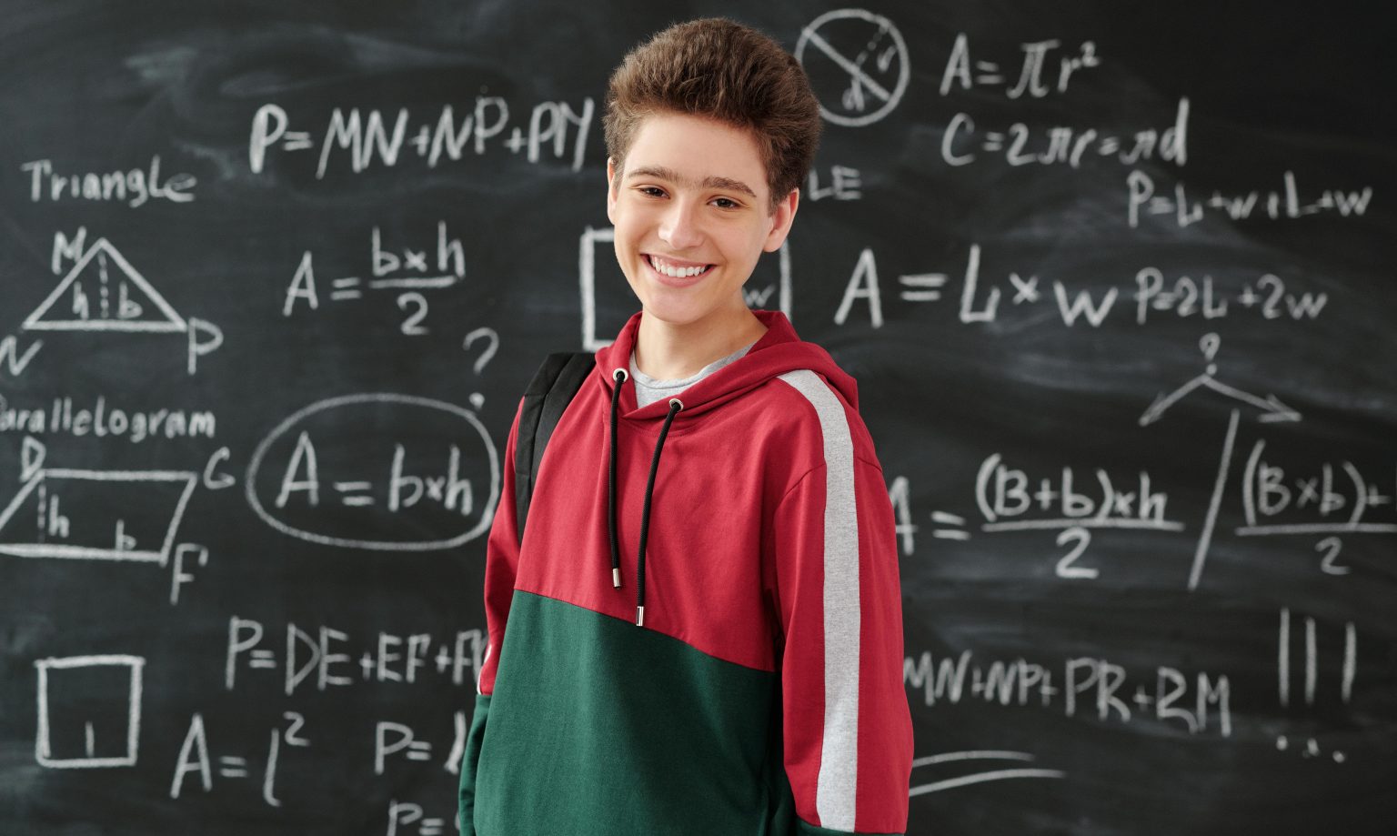 A young smiling boy stands in front of a blackboard scribbled with formulae