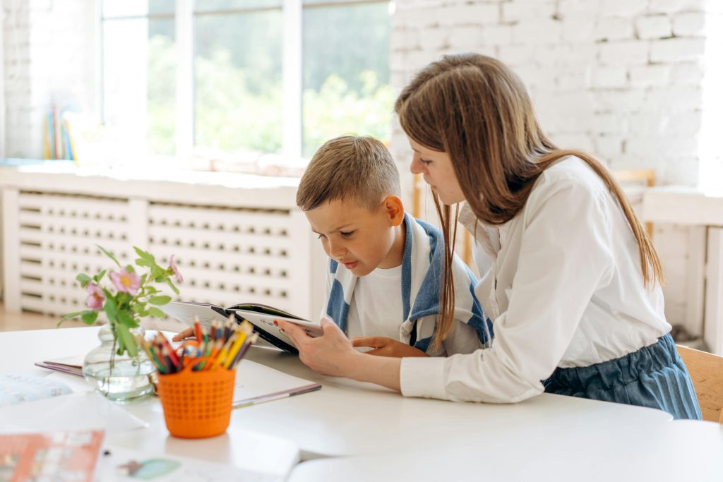 A mother with her child who has a book in his hand on a study desk which has color pencils and a plant