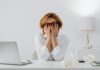 Overthinking Everything? 3 Simple Ways to Force-Quit Your Brain Stressed woman wearing glasses covering her face with hands at a desk next to a laptop and crumpled paper while overthinking