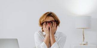 Overthinking Everything? 3 Simple Ways to Force-Quit Your Brain Stressed woman wearing glasses covering her face with hands at a desk next to a laptop and crumpled paper while overthinking