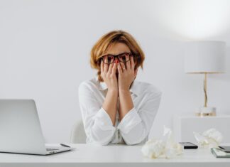 Overthinking Everything? 3 Simple Ways to Force-Quit Your Brain Stressed woman wearing glasses covering her face with hands at a desk next to a laptop and crumpled paper while overthinking