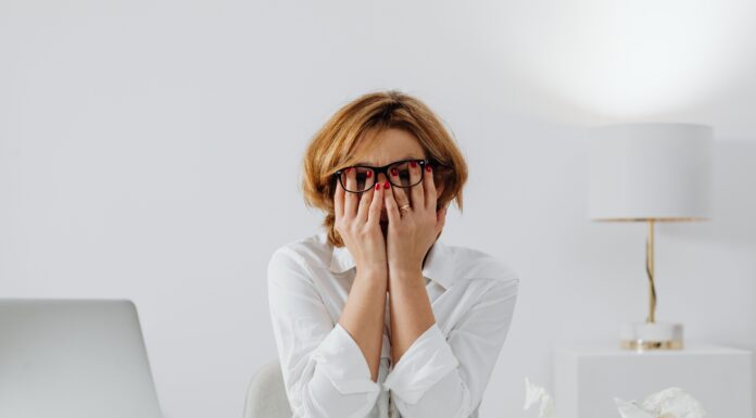 Overthinking Everything? 3 Simple Ways to Force-Quit Your Brain Stressed woman wearing glasses covering her face with hands at a desk next to a laptop and crumpled paper while overthinking