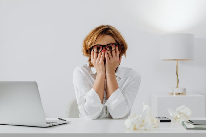how-to-stop-overthinking-stressed-woman-desk Stressed woman wearing glasses covering her face with hands at a desk next to a laptop and crumpled paper while overthinking