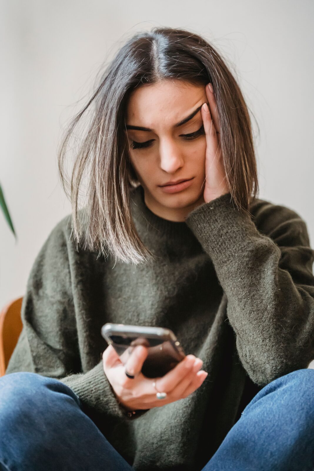 whatsapp-overthinking-blue-ticks.jpg A young woman sitting on a couch, looking down at her glowing smartphone screen with a subtle expression of sadness and anxiety, waiting for a reply.