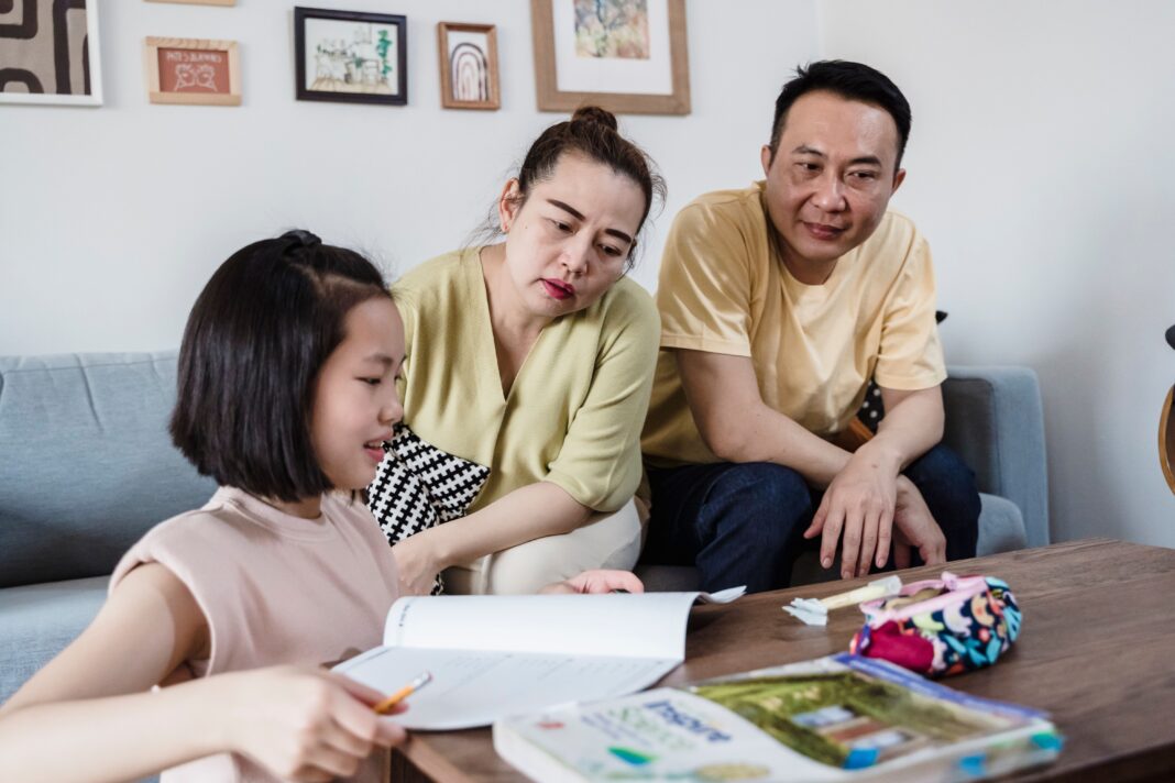 Building Critical Thinking: How Parents Can Help Kids Rely Less on AI for Homework An Asian mother and father attentively watching their young daughter work on a physical workbook at a coffee table with a pencil, encouraging offline problem-solving and focus without digital devices.