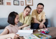 Modern Parenting: 5 Creative Ways Parents Can Break AI Dependency in Students An Asian mother and father attentively watching their young daughter work on a physical workbook at a coffee table with a pencil, encouraging offline problem-solving and focus without digital devices.