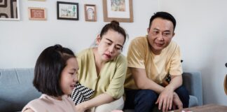 Modern Parenting: 5 Creative Ways Parents Can Break AI Dependency in Students An Asian mother and father attentively watching their young daughter work on a physical workbook at a coffee table with a pencil, encouraging offline problem-solving and focus without digital devices.
