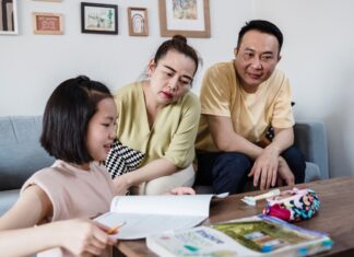 Modern Parenting: 5 Creative Ways Parents Can Break AI Dependency in Students An Asian mother and father attentively watching their young daughter work on a physical workbook at a coffee table with a pencil, encouraging offline problem-solving and focus without digital devices.