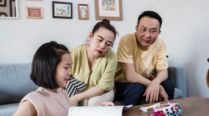 Modern Parenting: 5 Creative Ways Parents Can Break AI Dependency in Students An Asian mother and father attentively watching their young daughter work on a physical workbook at a coffee table with a pencil, encouraging offline problem-solving and focus without digital devices.