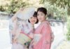 Ancient Wisdom – 10 Japanese Micro-Habits to Simplify Your Everyday Life Two smiling young women dressed in traditional pastel pink and floral Japanese kimonos, one holding up an open folding fan outdoors