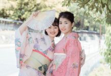 Ancient Wisdom – 10 Japanese Micro-Habits to Simplify Your Everyday Life Two smiling young women dressed in traditional pastel pink and floral Japanese kimonos, one holding up an open folding fan outdoors