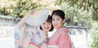 Ancient Wisdom – 10 Japanese Micro-Habits to Simplify Your Everyday Life Two smiling young women dressed in traditional pastel pink and floral Japanese kimonos, one holding up an open folding fan outdoors