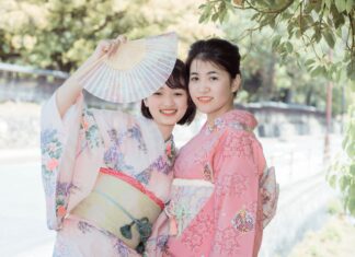 Ancient Wisdom – 10 Japanese Micro-Habits to Simplify Your Everyday Life Two smiling young women dressed in traditional pastel pink and floral Japanese kimonos, one holding up an open folding fan outdoors