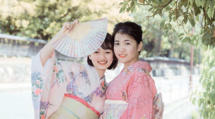 Ancient Wisdom – 10 Japanese Micro-Habits to Simplify Your Everyday Life Two smiling young women dressed in traditional pastel pink and floral Japanese kimonos, one holding up an open folding fan outdoors