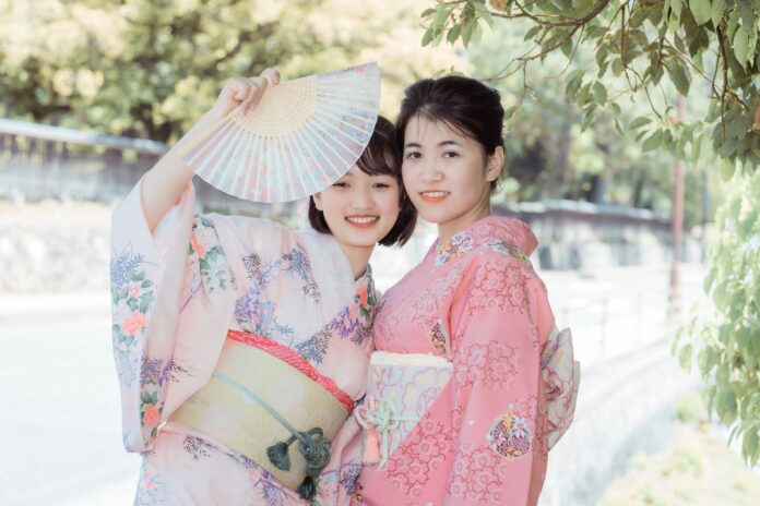 Two smiling young women dressed in traditional pastel pink and floral Japanese kimonos, one holding up an open folding fan outdoors
